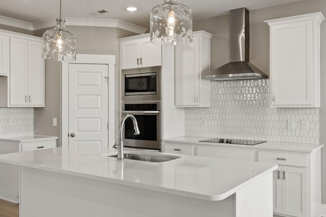 kitchen with wall chimney exhaust hood, appliances with stainless steel finishes, an island with sink, pendant lighting, and white cabinets