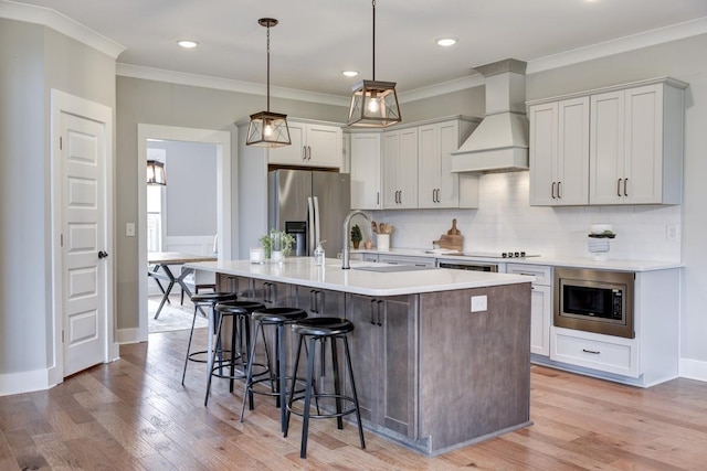 kitchen with pendant lighting, appliances with stainless steel finishes, white cabinetry, a kitchen island with sink, and custom exhaust hood