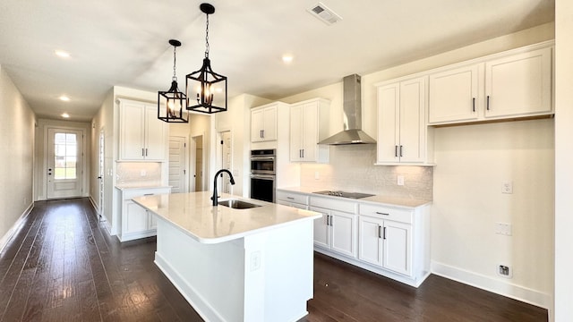 kitchen with sink, white cabinetry, a center island with sink, double oven, and wall chimney range hood