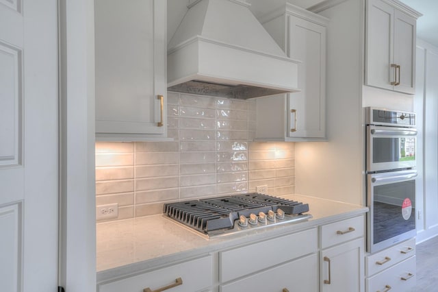 kitchen with appliances with stainless steel finishes, premium range hood, tasteful backsplash, light stone counters, and white cabinets