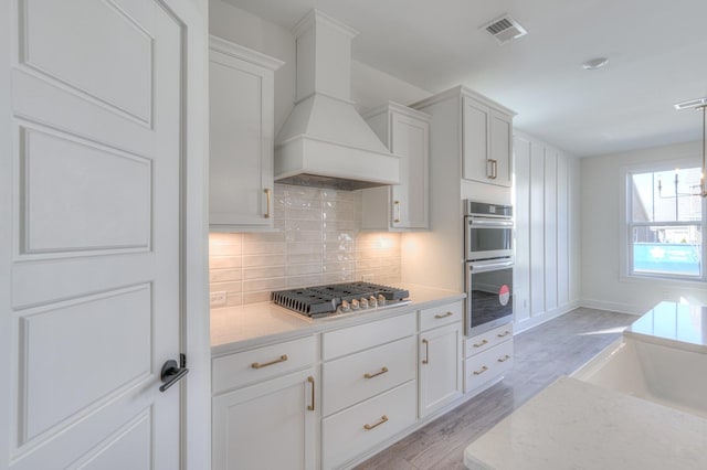 kitchen featuring premium range hood, white cabinets, backsplash, stainless steel appliances, and light stone countertops