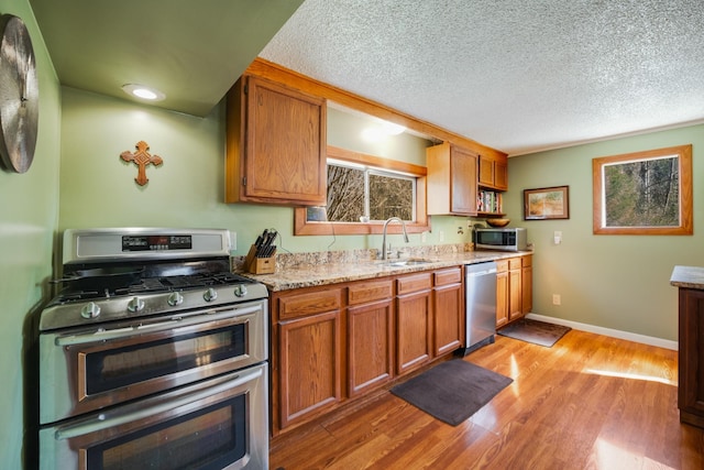 kitchen with sink, a textured ceiling, appliances with stainless steel finishes, light stone countertops, and light hardwood / wood-style floors