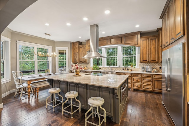 kitchen featuring island range hood, brown cabinetry, light stone counters, a center island, and stainless steel built in fridge