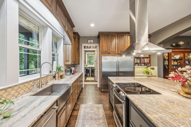 kitchen featuring island exhaust hood, appliances with stainless steel finishes, washing machine and dryer, a sink, and light stone countertops