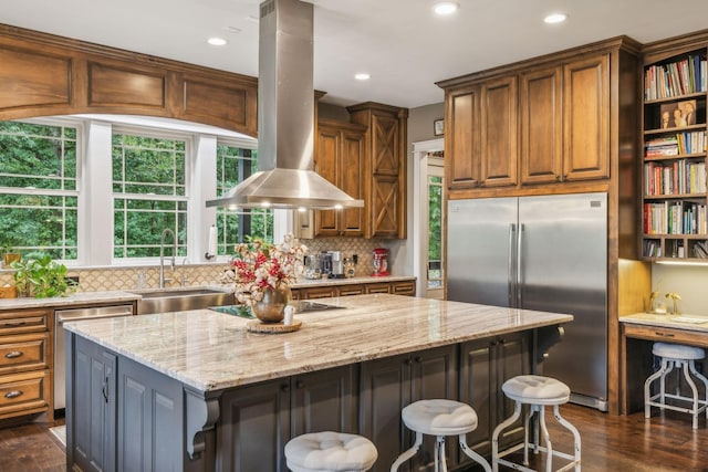 kitchen with stainless steel appliances, a sink, a kitchen island, light stone countertops, and island exhaust hood