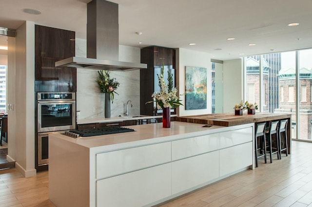 kitchen with light wood-type flooring, backsplash, double oven, island range hood, and light countertops