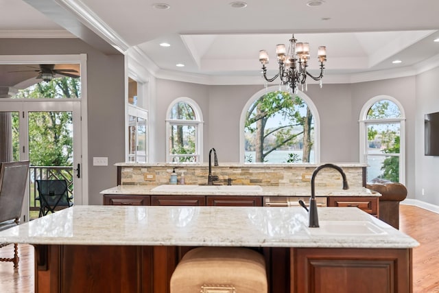 kitchen with sink, a center island with sink, a raised ceiling, light stone countertops, and decorative backsplash