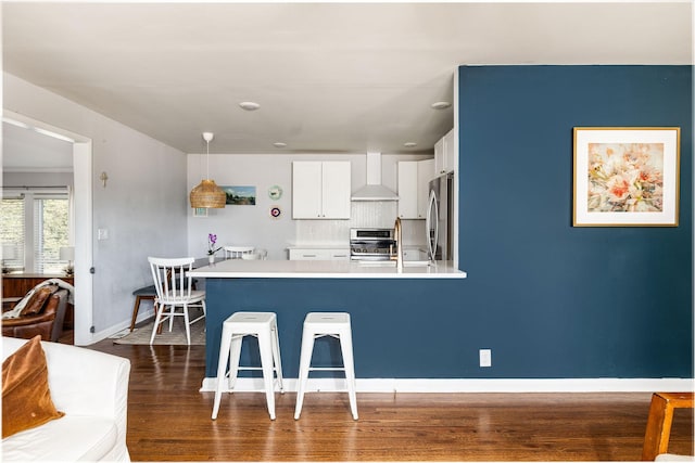 kitchen with white cabinetry, hanging light fixtures, appliances with stainless steel finishes, a kitchen breakfast bar, and wall chimney range hood