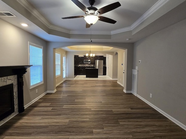 unfurnished living room with ceiling fan with notable chandelier, dark hardwood / wood-style floors, ornamental molding, a stone fireplace, and a raised ceiling