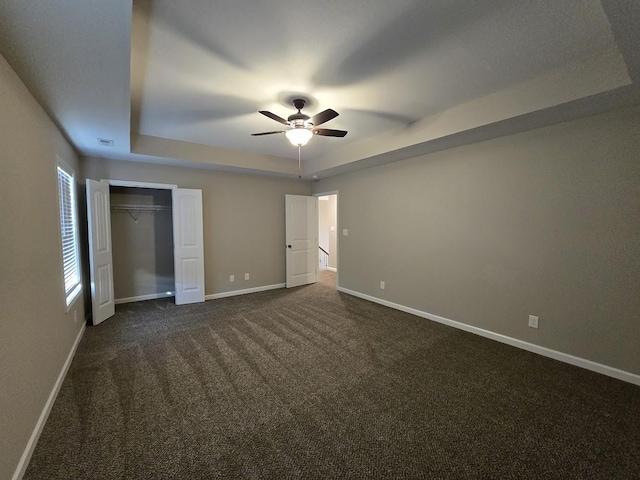 unfurnished bedroom featuring a raised ceiling, ceiling fan, dark carpet, and a closet