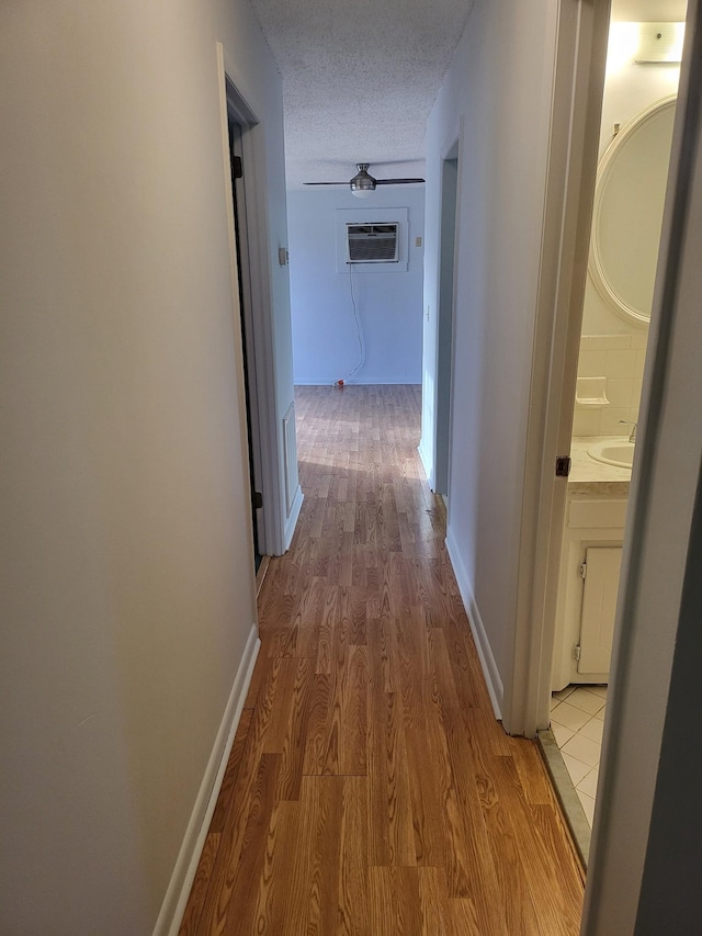 hall with an AC wall unit, a textured ceiling, baseboards, and light wood finished floors