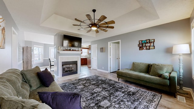 living room with a raised ceiling, a tiled fireplace, and ceiling fan