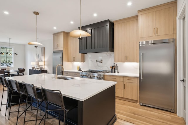 kitchen featuring premium appliances, decorative light fixtures, light brown cabinets, and a center island with sink