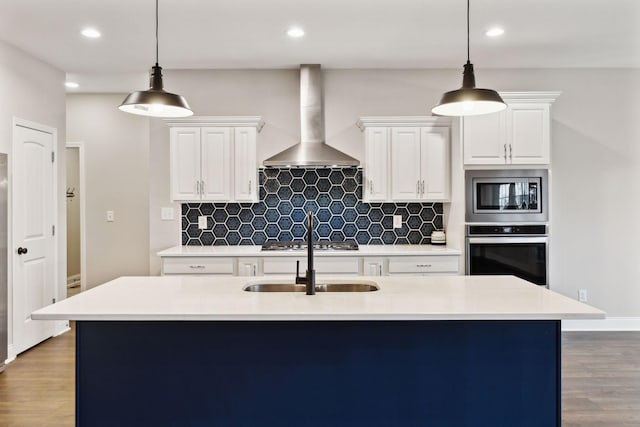 kitchen featuring appliances with stainless steel finishes, white cabinetry, hanging light fixtures, a center island with sink, and wall chimney exhaust hood