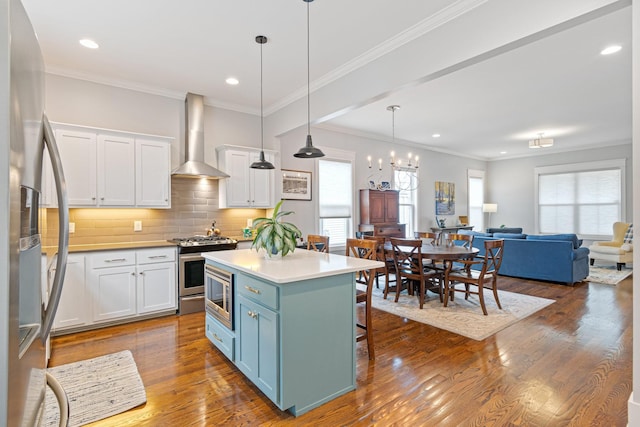 kitchen with wall chimney range hood, stainless steel appliances, hanging light fixtures, and white cabinets