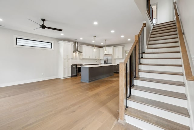 kitchen with wall chimney range hood, appliances with stainless steel finishes, hanging light fixtures, an island with sink, and white cabinets