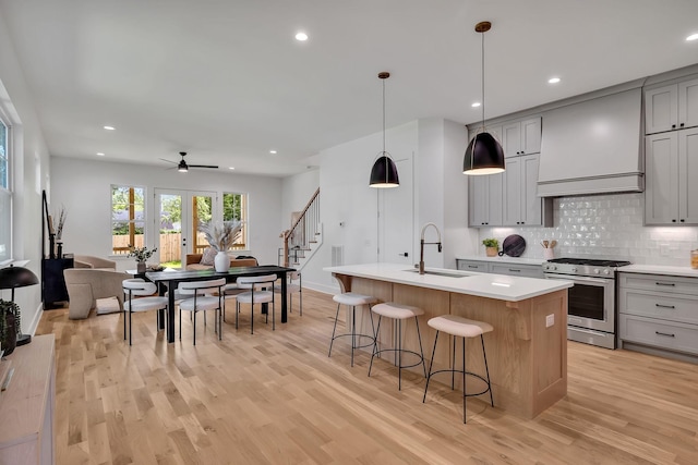 kitchen featuring sink, custom exhaust hood, stainless steel gas range oven, gray cabinets, and a kitchen island with sink