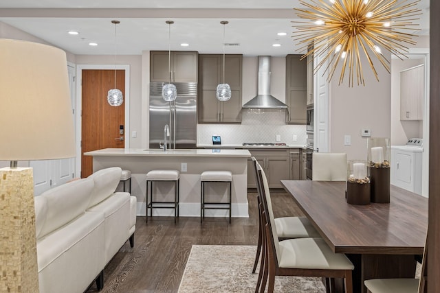 kitchen with gray cabinets, dark hardwood / wood-style floors, a kitchen island with sink, built in appliances, and wall chimney range hood