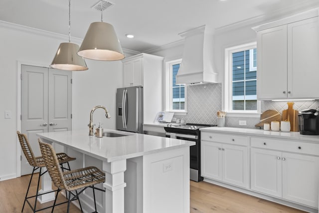 kitchen with custom exhaust hood, white cabinetry, decorative light fixtures, appliances with stainless steel finishes, and an island with sink