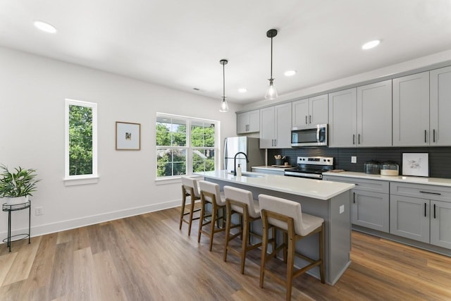 kitchen with sink, a kitchen island with sink, gray cabinetry, stainless steel appliances, and decorative light fixtures