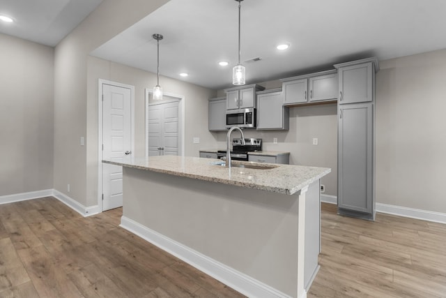 kitchen featuring sink, hanging light fixtures, stainless steel appliances, light stone countertops, and a kitchen island with sink