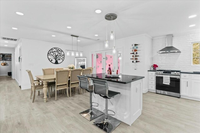 kitchen with stainless steel gas range, hanging light fixtures, a kitchen breakfast bar, wall chimney range hood, and white cabinets