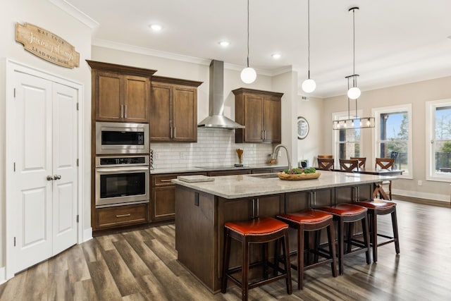 kitchen with an island with sink, hanging light fixtures, stainless steel appliances, sink, and wall chimney exhaust hood