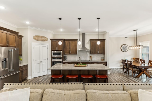 kitchen featuring a center island with sink, decorative light fixtures, appliances with stainless steel finishes, wall chimney range hood, and a kitchen bar