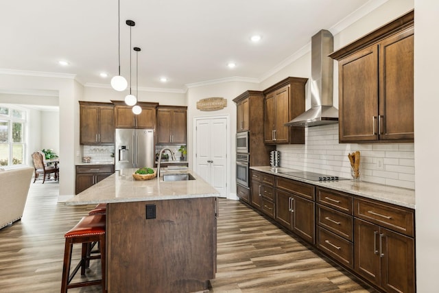 kitchen featuring a center island with sink, sink, stainless steel appliances, wall chimney exhaust hood, and hanging light fixtures