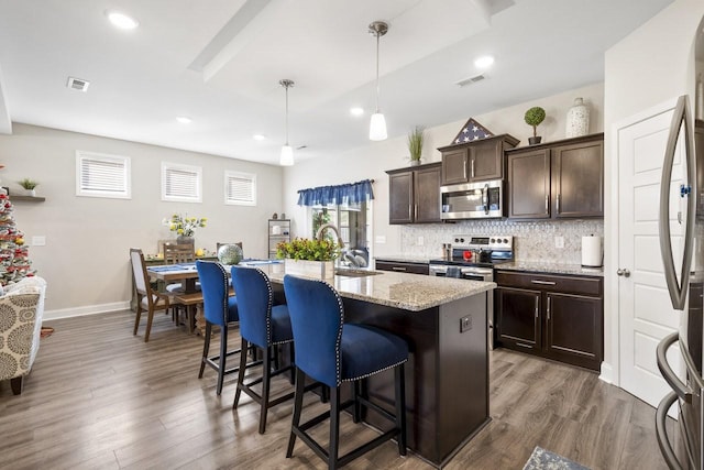 kitchen featuring appliances with stainless steel finishes, hanging light fixtures, dark brown cabinetry, light stone countertops, and a center island with sink