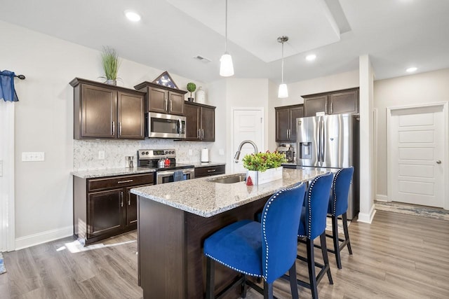 kitchen with sink, a kitchen island with sink, hanging light fixtures, stainless steel appliances, and light stone counters