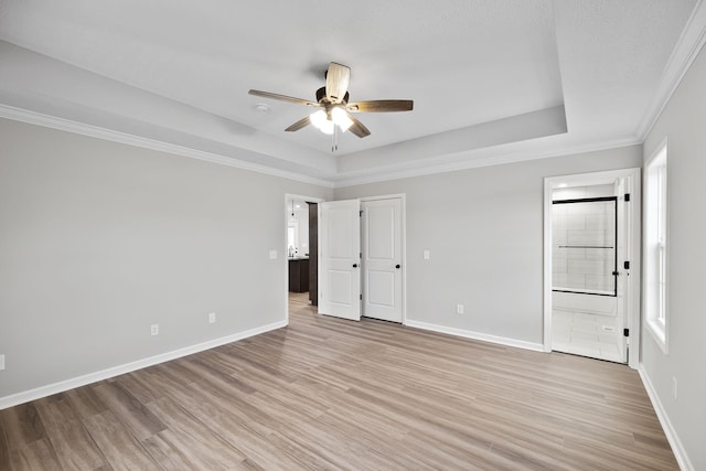 unfurnished bedroom featuring ensuite bath, ceiling fan, a tray ceiling, ornamental molding, and light wood-type flooring