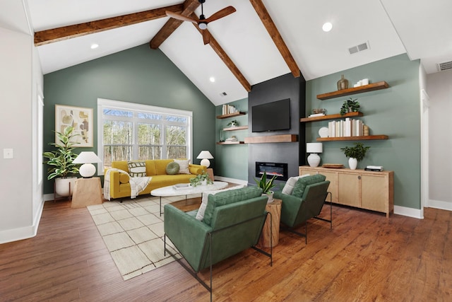 living area with high vaulted ceiling, wood finished floors, a ceiling fan, beam ceiling, and a glass covered fireplace