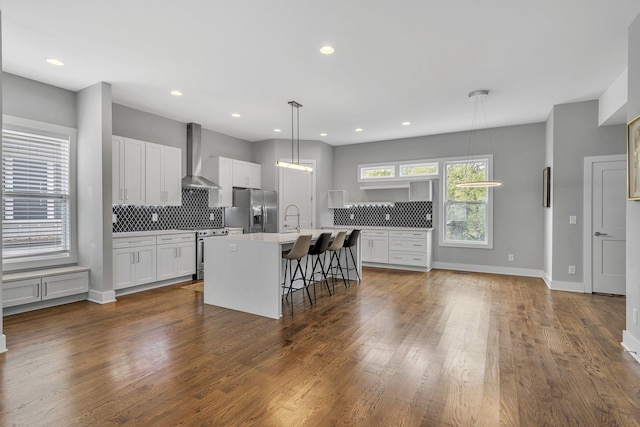 kitchen with a center island with sink, wall chimney exhaust hood, stainless steel appliances, pendant lighting, and white cabinetry