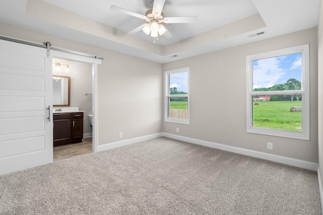 unfurnished bedroom featuring connected bathroom, ceiling fan, light carpet, a tray ceiling, and a barn door