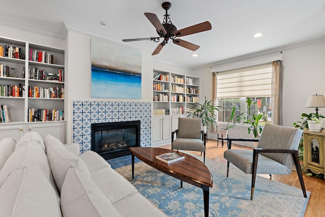 living room featuring light wood-type flooring, a tile fireplace, ceiling fan, built in features, and ornamental molding