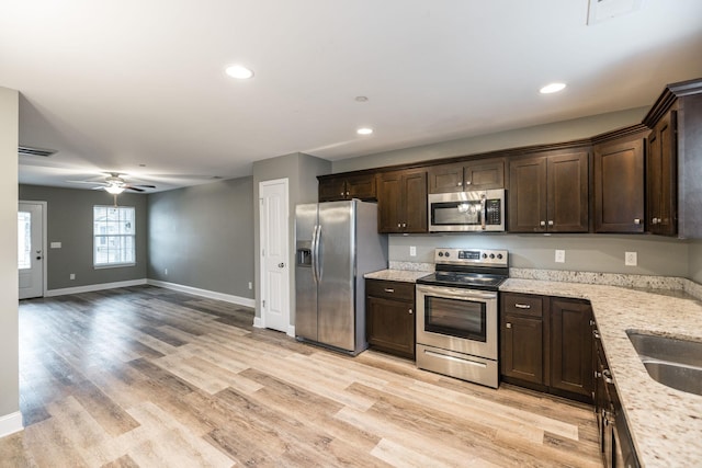 kitchen with light wood-type flooring, stainless steel appliances, dark brown cabinetry, ceiling fan, and light stone counters