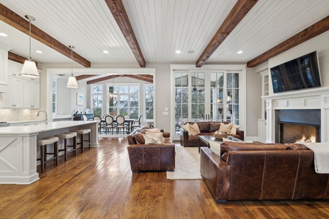 living area featuring a warm lit fireplace, baseboards, dark wood-type flooring, beam ceiling, and recessed lighting