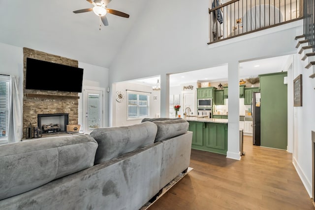 living room with high vaulted ceiling, a fireplace, light wood finished floors, and ceiling fan with notable chandelier