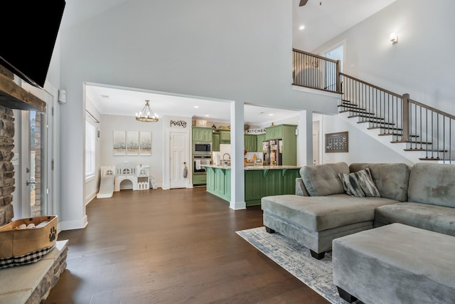 living area featuring baseboards, dark wood finished floors, a towering ceiling, stairs, and ceiling fan with notable chandelier