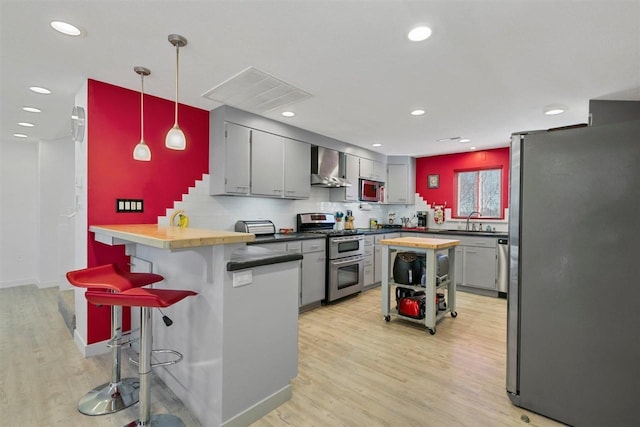 kitchen with butcher block countertops, decorative light fixtures, appliances with stainless steel finishes, wall chimney range hood, and a kitchen breakfast bar