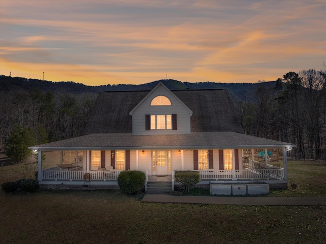 farmhouse inspired home with covered porch, a shingled roof, a lawn, and a wooded view