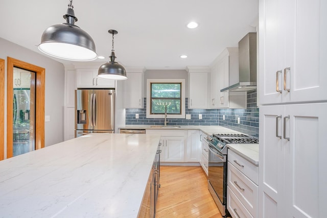 kitchen featuring stainless steel refrigerator with ice dispenser, range with gas stovetop, wall chimney range hood, white cabinets, and hanging light fixtures