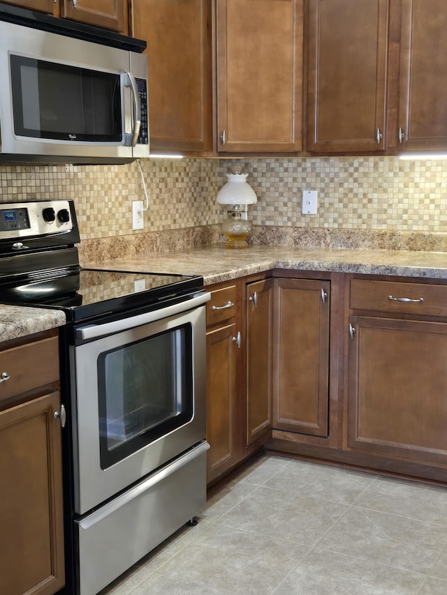 kitchen with light tile patterned flooring, appliances with stainless steel finishes, backsplash, light stone countertops, and brown cabinetry