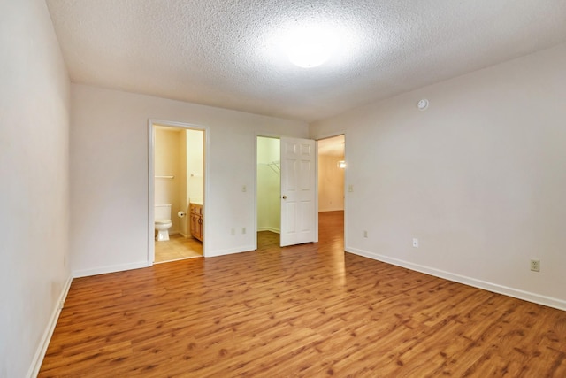 unfurnished bedroom featuring a walk in closet, ensuite bathroom, a textured ceiling, light wood-type flooring, and baseboards