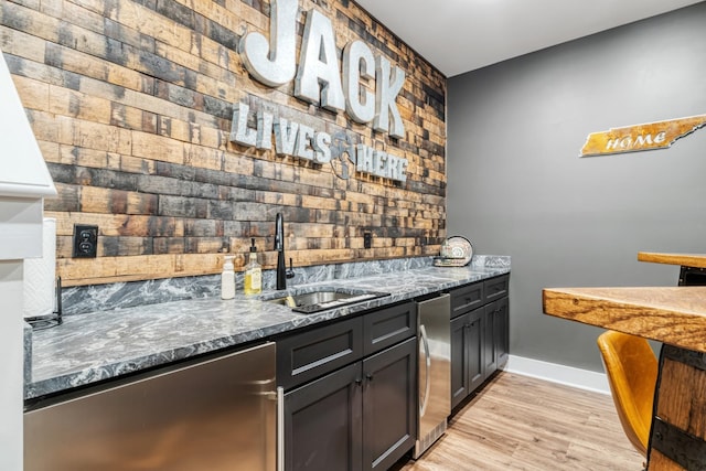 bar with stainless steel dishwasher, light hardwood / wood-style flooring, sink, and dark stone countertops