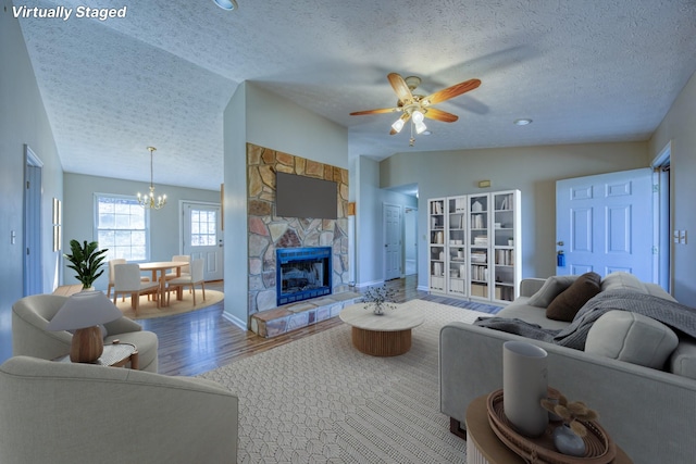 living room featuring vaulted ceiling, a stone fireplace, a textured ceiling, wood finished floors, and ceiling fan with notable chandelier