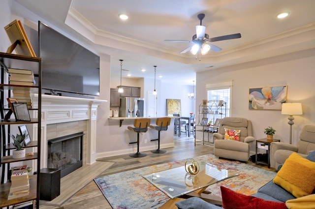 living room featuring light wood-type flooring, a tray ceiling, a tiled fireplace, ceiling fan, and crown molding
