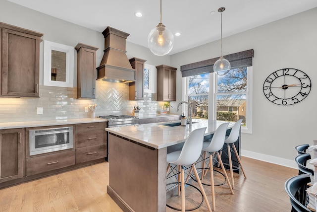 kitchen with light wood-type flooring, custom range hood, pendant lighting, sink, and a kitchen island with sink