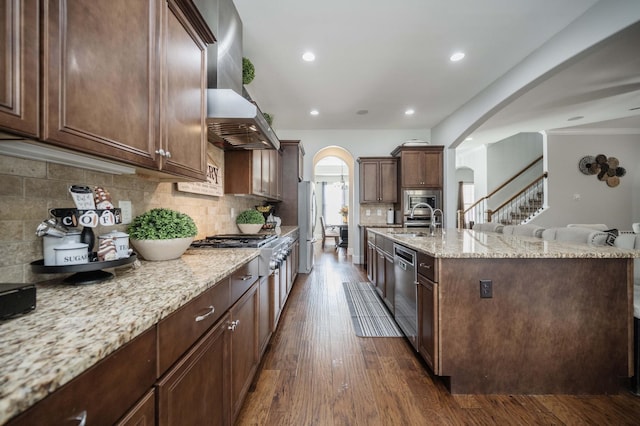 kitchen featuring arched walkways, stainless steel appliances, dark wood-type flooring, a sink, and wall chimney range hood